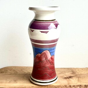 May include: A white ceramic vase with a geometric design in shades of purple, blue, and red. The vase has a wide mouth and a narrow base.