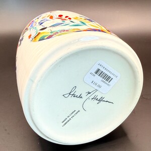 May include: Cream-colored ceramic container with a colorful floral design. The container has vertical ribbed detailing and a slightly flared top. The bottom has a sticker with the brand name and a handwritten signature.