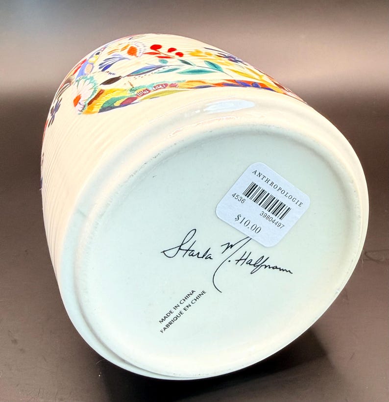 May include: Cream-colored ceramic container with a colorful floral design. The container has vertical ribbed detailing and a slightly flared top. The bottom has a sticker with the brand name and a handwritten signature.