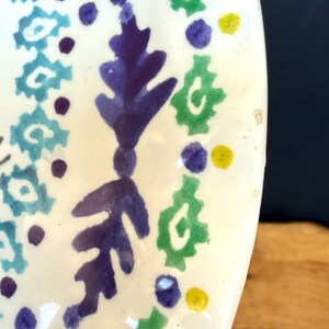 Puede incluir: Fragmento de un plato de cer&aacute;mica blanca con un dise&ntilde;o pintado a mano. El dise&ntilde;o presenta patrones florales y geom&eacute;tricos azules, verdes, morados y amarillos. El plato tiene un acabado brillante y una forma irregular.