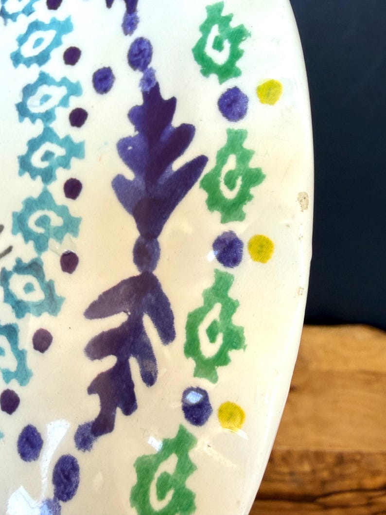 Puede incluir: Fragmento de un plato de cer&aacute;mica blanca con un dise&ntilde;o pintado a mano. El dise&ntilde;o presenta patrones florales y geom&eacute;tricos azules, verdes, morados y amarillos. El plato tiene un acabado brillante y una forma irregular.