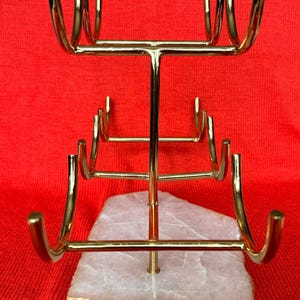 May include: A gold-coloured metal jewellery display stand with multiple curved arms for hanging necklaces or bracelets. The stand is mounted on a white and orange stone base, set against a red background. The stand is designed to showcase jewellery.
