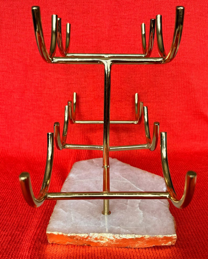 May include: A gold-coloured metal jewellery display stand with multiple curved arms for hanging necklaces or bracelets. The stand is mounted on a white and orange stone base, set against a red background. The stand is designed to showcase jewellery.