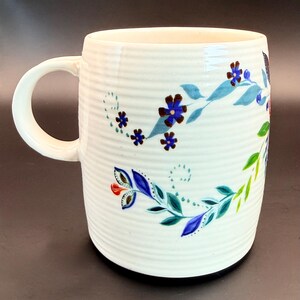 May include: White ceramic mug with a ribbed texture and a curved handle. The mug is decorated with a floral design featuring blue, green, and red flowers and leaves. The design wraps around the mug.