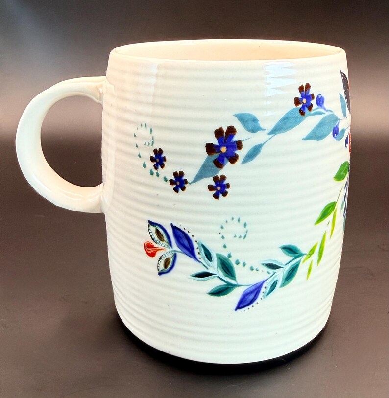 May include: White ceramic mug with a ribbed texture and a curved handle. The mug is decorated with a floral design featuring blue, green, and red flowers and leaves. The design wraps around the mug.