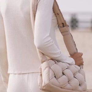K&ouml;nnte beinhalten: Eine beige gewebte Umh&auml;ngetasche mit breitem Riemen. Die Tasche ist rechteckig mit einem gepolsterten, gewebten Design. Der Riemen ist ebenfalls beige und scheint verstellbar zu sein. Die Person tr&auml;gt ein cremefarbenes Rippstrick-Oberteil und eine Hose.