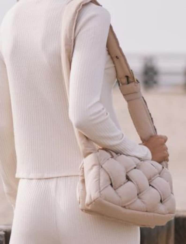 K&ouml;nnte beinhalten: Eine beige gewebte Umh&auml;ngetasche mit breitem Riemen. Die Tasche ist rechteckig mit einem gepolsterten, gewebten Design. Der Riemen ist ebenfalls beige und scheint verstellbar zu sein. Die Person tr&auml;gt ein cremefarbenes Rippstrick-Oberteil und eine Hose.