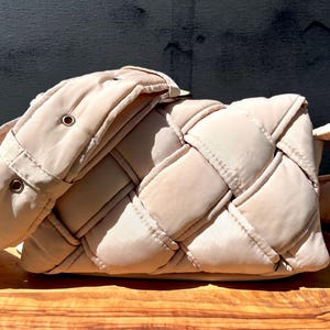 K&ouml;nnte beinhalten: Beige gewebte Handtasche mit gepolstertem Design. Die Tasche hat ein gewebtes Muster auf dem Hauptteil und einen passenden gepolsterten Riemen. Die Tasche ist rechteckig mit abgerundeten Ecken.