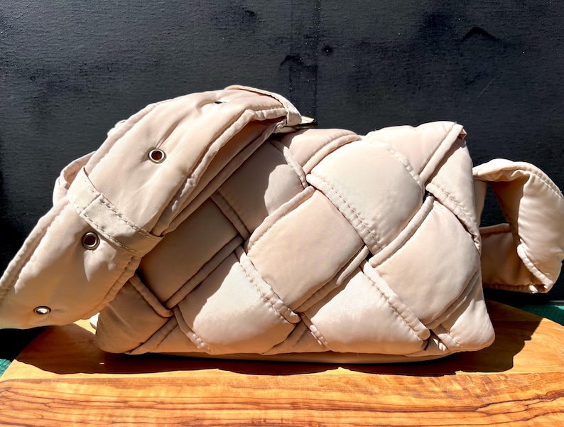 K&ouml;nnte beinhalten: Beige gewebte Handtasche mit gepolstertem Design. Die Tasche hat ein gewebtes Muster auf dem Hauptteil und einen passenden gepolsterten Riemen. Die Tasche ist rechteckig mit abgerundeten Ecken.