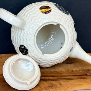 May include: White ceramic teapot with a textured, woven-like pattern. The teapot features a spout, handle, and a lid. It is decorated with black, yellow, and red circular designs. The lid is off to the side, revealing the interior.
