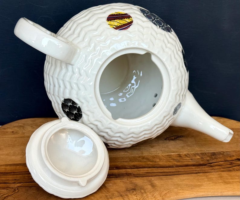 May include: White ceramic teapot with a textured, woven-like pattern. The teapot features a spout, handle, and a lid. It is decorated with black, yellow, and red circular designs. The lid is off to the side, revealing the interior.