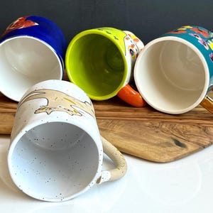 May include: Four ceramic mugs with various designs. One is blue with a red floral pattern, another is lime green, a third is white with a speckled pattern and a cartoon animal, and the last is teal with colourful flowers and a gold handle.