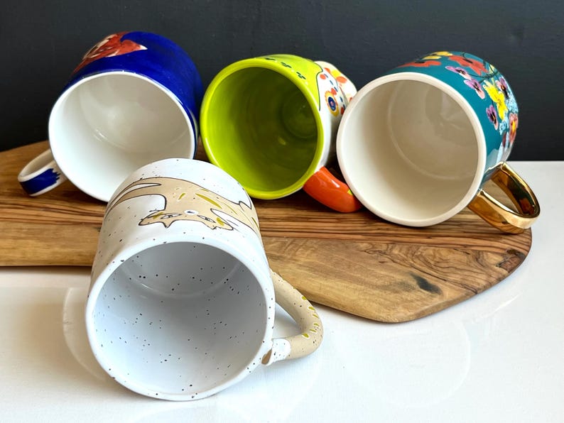 May include: Four ceramic mugs with various designs. One is blue with a red floral pattern, another is lime green, a third is white with a speckled pattern and a cartoon animal, and the last is teal with colourful flowers and a gold handle.