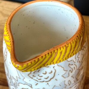 May include: A ceramic pitcher with a white interior and a patterned exterior. The rim is accented with a yellow and brown design. The pitcher has a handle and a spout for pouring.