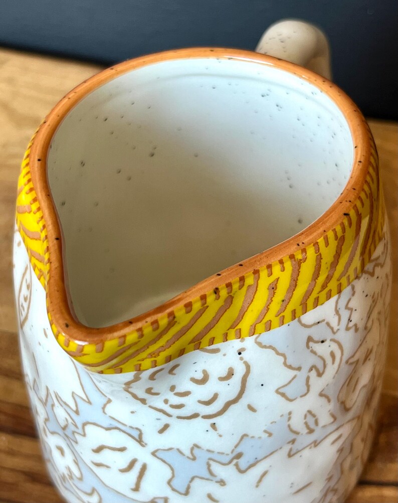 May include: A ceramic pitcher with a white interior and a patterned exterior. The rim is accented with a yellow and brown design. The pitcher has a handle and a spout for pouring.