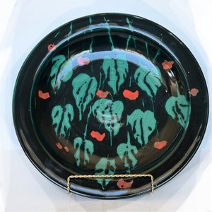 May include: A black ceramic plate with a green and red floral design. The plate has a raised rim and is decorated with a pattern of leaves and flowers.