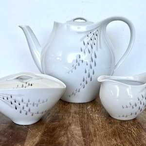 May include: A white teapot, creamer, and sugar bowl with a gray floral design. The teapot has a curved spout and a handle on the side. The creamer and sugar bowl have a rounded shape.