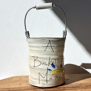 May include: A ceramic bucket with a wire handle and a white ceramic handle. The bucket is painted with a cream and gray striped pattern and the words "A Bucket of Maze" in black script. Blue and yellow accents are also present.