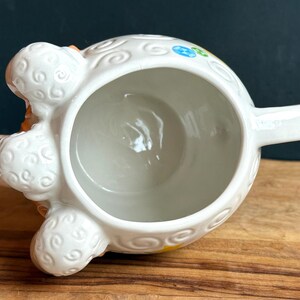 May include: White ceramic sheep-shaped creamer with a handle. The creamer features a detailed design with curly wool texture and colorful floral accents. The sheep's face is peach-colored with closed eyes. The creamer sits on a wooden surface.