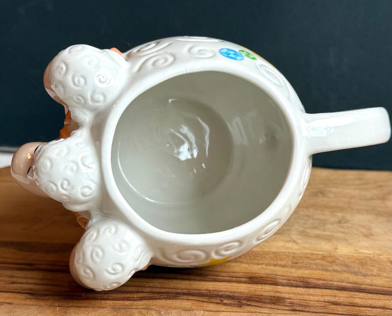 May include: White ceramic sheep-shaped creamer with a handle. The creamer features a detailed design with curly wool texture and colorful floral accents. The sheep's face is peach-colored with closed eyes. The creamer sits on a wooden surface.