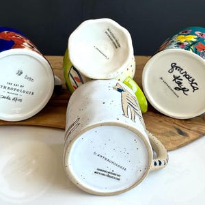 May include: Four ceramic mugs with distinctive designs are shown. One mug has a blue background with a bird, another features a cat illustration, and a third has a floral pattern. The mugs are from Anthropologie.