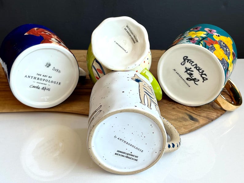 May include: Four ceramic mugs with distinctive designs are shown. One mug has a blue background with a bird, another features a cat illustration, and a third has a floral pattern. The mugs are from Anthropologie.