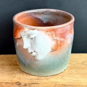 May include: A handmade ceramic cup with a sculpted face on the side. The cup features a blend of colors, including orange, blue, and green. The face is a light color, contrasting with the darker hues of the cup.