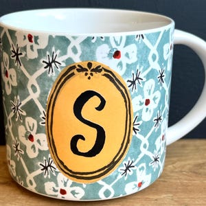 Pu&ograve; includere: Tazza in ceramica bianca con un motivo floreale turchese e la lettera "S" in un ovale giallo. La tazza ha un manico bianco e un design decorativo con fiori bianchi e stelle nere.