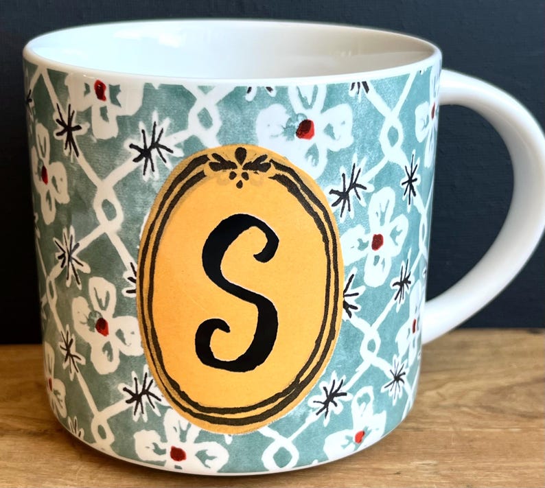 Pu&ograve; includere: Tazza in ceramica bianca con un motivo floreale turchese e la lettera "S" in un ovale giallo. La tazza ha un manico bianco e un design decorativo con fiori bianchi e stelle nere.