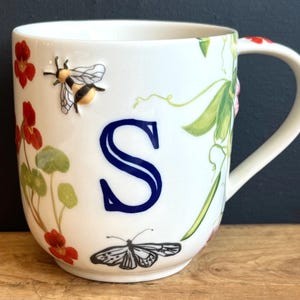 Pu&ograve; includere: Tazza in ceramica bianca con una grande lettera "S" blu e illustrazioni floreali e di insetti. La tazza presenta un'ape, una farfalla, fiori rossi e foglie verdi. Il manico ha un accento floreale rosso.