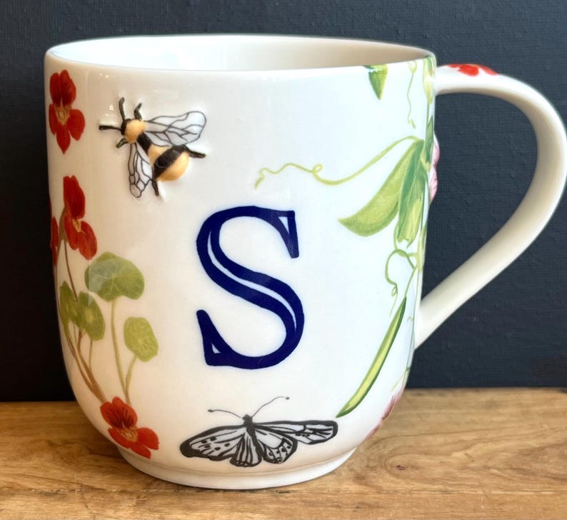 Pu&ograve; includere: Tazza in ceramica bianca con una grande lettera "S" blu e illustrazioni floreali e di insetti. La tazza presenta un'ape, una farfalla, fiori rossi e foglie verdi. Il manico ha un accento floreale rosso.