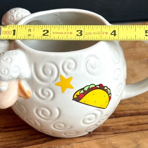May include: White ceramic sheep-shaped mug with a handle. The mug features a yellow star and a taco graphic. The mug is approximately 4 inches in diameter.