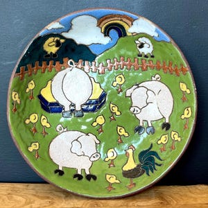 May include: A hand-painted ceramic plate with a whimsical farm scene. The plate features three white pigs, yellow chicks, and sheep on a green field, with a rainbow and clouds in the background. The rim is brown.