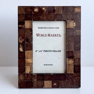May include: A rectangular photo frame featuring a mosaic border of small, square, dark brown and tan tiles. The center insert displays the text: MADE EXCLUSIVELY FOR WORLD MARKET, 4" x 6" PHOTO FRAME, MADE IN INDIA. The frame has a vertical orientation.