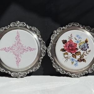 May include: Two decorative silver-toned trays with white ceramic centers. One features a pink floral cross design, the other a red rose and blue floral arrangement. The trays have ornate, scalloped edges.