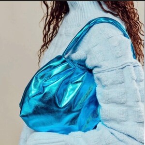 May include: A bright blue, metallic-looking handbag with a gathered design. The bag has a matching strap and is held over the shoulder. The bag's shiny surface reflects light, creating a vibrant appearance. The bag is a fashion accessory.