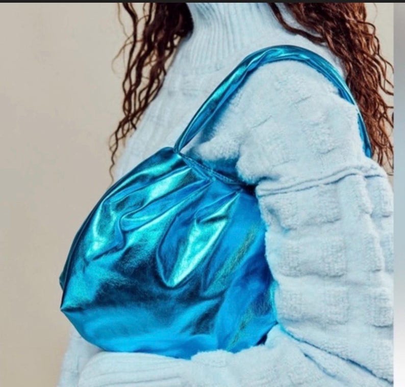 May include: A bright blue, metallic-looking handbag with a gathered design. The bag has a matching strap and is held over the shoulder. The bag's shiny surface reflects light, creating a vibrant appearance. The bag is a fashion accessory.