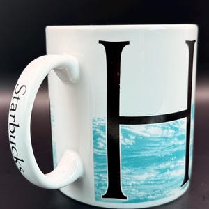 May include: White ceramic mug with a large black letter "H" and a turquoise wave design. The handle is white with the word "Starbucks" printed in black. The mug is set against a dark background.