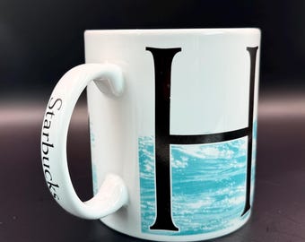 May include: White ceramic mug with a large black letter "H" and a turquoise wave design. The handle is white with the word "Starbucks" printed in black. The mug is set against a dark background.