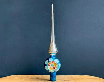 May include: A vintage Christmas tree topper with a silver spire and a round, blue base. The base features a colorful floral design with red, yellow, and green accents. The topper is made of glass and has a classic, retro aesthetic.