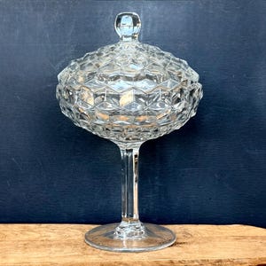May include: A clear, vintage glass candy dish with a lid. The dish has a diamond-cut pattern and sits on a tall, slender stem. The lid has a small, round handle. The dish is displayed on a wooden surface against a dark blue background.