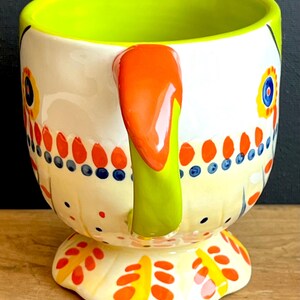 May include: A ceramic mug with a white base, decorated with colourful floral and dot patterns. The interior is bright green, with an orange handle and a yellow footed base. The mug has a whimsical, artistic design.