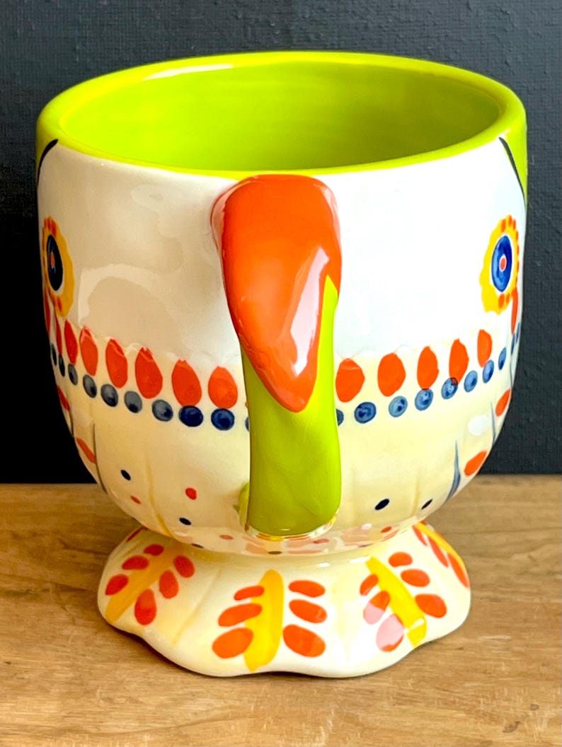 May include: A ceramic mug with a white base, decorated with colourful floral and dot patterns. The interior is bright green, with an orange handle and a yellow footed base. The mug has a whimsical, artistic design.