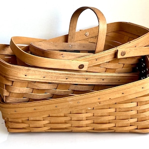 May include: Three woven wooden baskets of varying sizes, stacked together. The largest basket is at the bottom, with a rectangular shape and a handle. The other two baskets are smaller, with handles and lids. The baskets are light brown.
