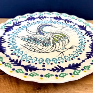 Puede incluir: Un plato de cer&aacute;mica blanco con borde festoneado, con un dise&ntilde;o de pavo real pintado a mano en tonos azules, verdes y negros. El plato tiene un borde decorativo con motivos florales y geom&eacute;tricos.