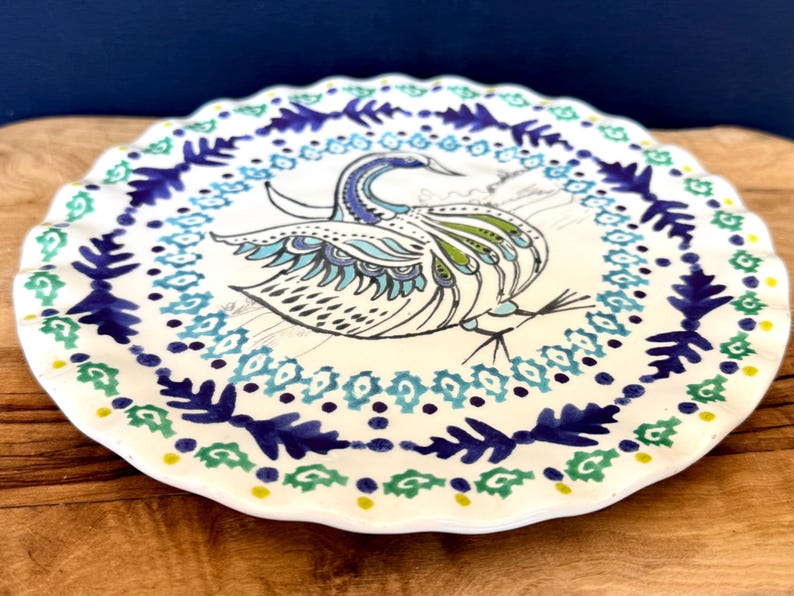 Puede incluir: Un plato de cer&aacute;mica blanco con borde festoneado, con un dise&ntilde;o de pavo real pintado a mano en tonos azules, verdes y negros. El plato tiene un borde decorativo con motivos florales y geom&eacute;tricos.
