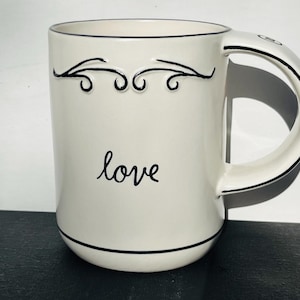 May include: White ceramic mug with a black outline and a black floral design. The mug has the word "love" written in black cursive on the front.