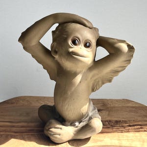 May include: A ceramic figurine of a monkey with large eyes and a playful expression. The monkey is sitting with its arms raised above its head, as if it is stretching or relaxing.