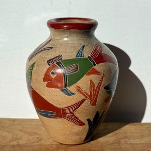 May include: A beige ceramic vase with a red rim and colorful fish designs. The fish are painted in red, green, blue, and black, with orange and black seaweed accents. The vase has a rounded body and a narrow neck.