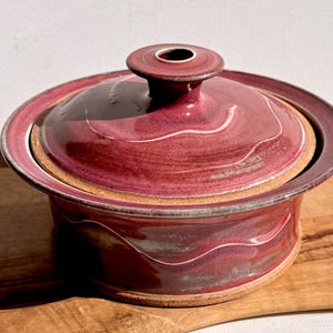 May include: A burgundy ceramic casserole dish with a lid. The dish has a round shape with a wide rim and a small handle on the lid. White wavy lines decorate the dish. The dish is sitting on a wooden cutting board.